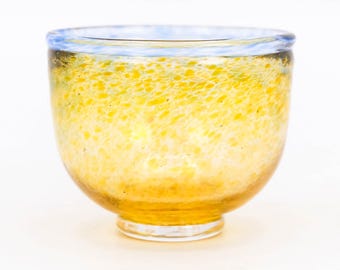 May include: A handcrafted glass bowl with a rounded shape. The bowl features a speckled yellow and orange design, with a light blue rim. The glass is transparent, allowing light to pass through. The bowl sits on a clear base.