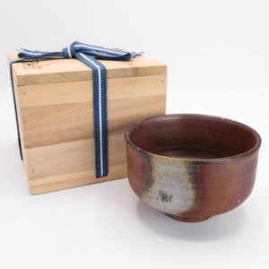 Puede incluir: Un cuenco de té de cerámica marrón y gris con una caja de madera atada con una cinta azul. La caja tiene caracteres japoneses en la parte superior.
