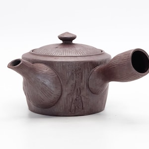 Könnte beinhalten: Eine braune Teekanne aus Keramik mit einer strukturierten Oberfläche und einem Ausguss in Form eines Bambusrohrs. Die Teekanne hat einen Deckel mit einem kleinen Knopf oben. Die Teekanne ist mit japanischen Schriftzeichen beschriftet.