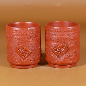 Puede incluir: Dos tazas de té de arcilla roja con superficies texturizadas ásperas y caracteres chinos inscritos en la parte delantera.