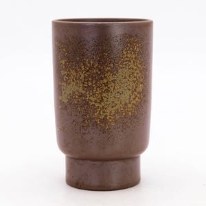 May include: A cylindrical, brown ceramic vase with a speckled, textured surface. The vase features a base and a speckled design in shades of brown and olive green. The vase is suitable for displaying flowers or as a decorative item.