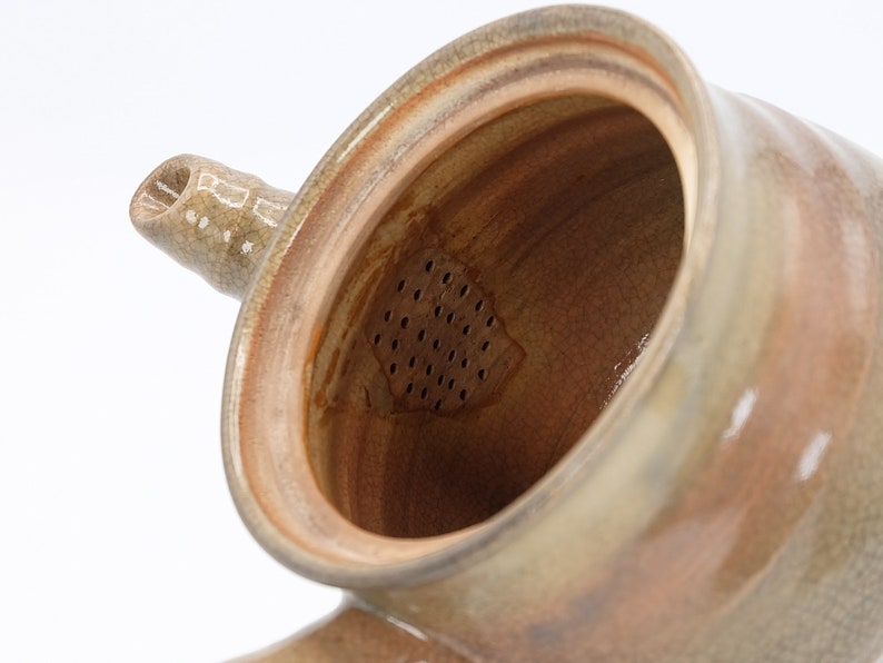 Może przedstawiać: Brązowy ceramiczny czajnik z wbudowanym sitkiem. Czajnik ma jasnobrązową glazurę z pękniętym wykończeniem. Sitko jest widoczne przez otw&oacute;r czajnika.
