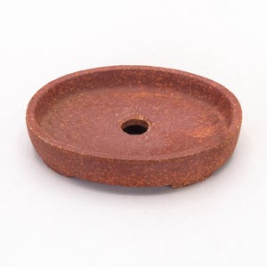 May include: A round, reddish-brown ceramic bonsai pot with a central drainage hole. The pot has a textured surface and a low, oval shape. The pot is designed for bonsai trees.