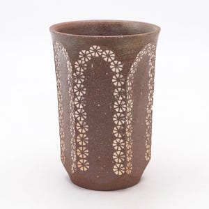 May include: A brown ceramic tumbler with a textured surface and a floral pattern. The tumbler features a series of white flower-shaped designs arranged in vertical rows and arches. The cup has a slightly flared rim.