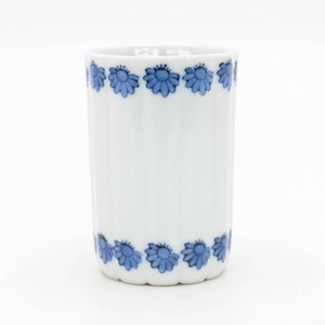 May include: A white ceramic tumbler with vertical ridges and a blue floral pattern around the top and bottom. The blue flowers have a stylized daisy design. The tumbler is cylindrical and stands upright against a white background.