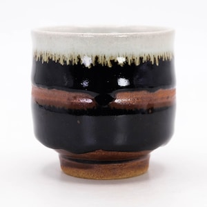 Pode incluir: Uma chávena de cerâmica com uma borda branca, uma faixa central preta e uma faixa castanha. A base é castanha. A chávena tem um acabamento brilhante e é provavelmente feita à mão. O design é simples e elegante, adequado para bebidas.