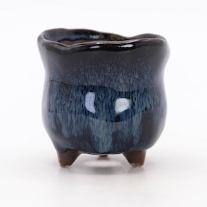 May include: A small, three-legged ceramic planter featuring a glossy, dark navy blue glaze with lighter blue, streaky vertical drip patterns. The rim has an irregular, wavy edge, and the three short, sturdy feet are a solid brown color.