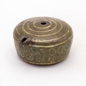 Może przedstawiać: Ręcznie wykonane ceramiczne naczynie z ciemno oliwkowozieloną glazurą. Na górze znajdują się koncentryczne okręgi, a na dolnej części wzór kwiatowy. Posiada dzióbek i mały otwór na górze.