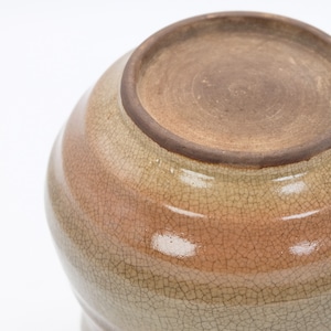 Może przedstawiać: Ceramiczny garnek z jasnobrązową glazurą i pękniętym wykończeniem. Garnek ma zaokrąglony kształt i szeroki otw&oacute;r.
