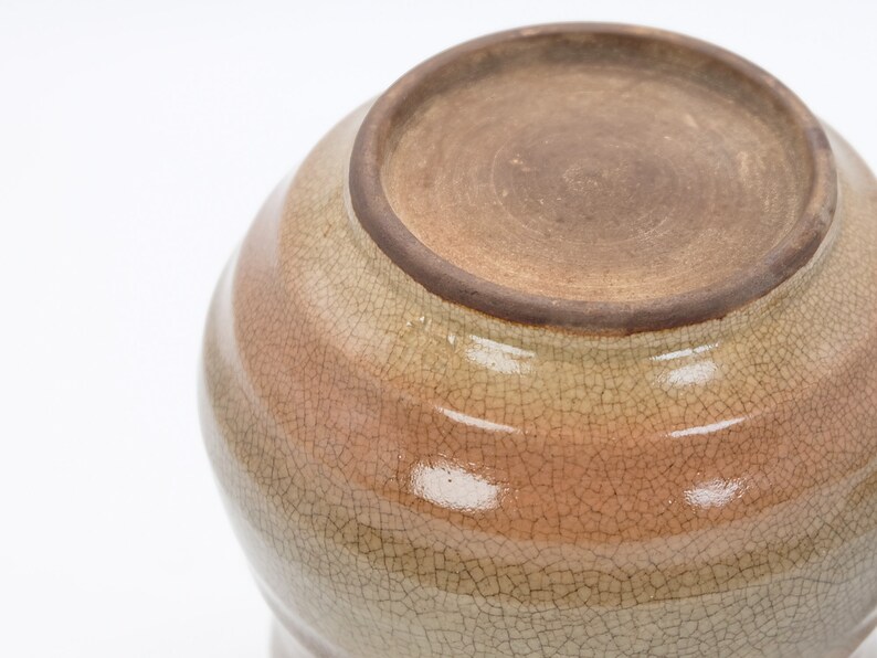 Może przedstawiać: Ceramiczny garnek z jasnobrązową glazurą i pękniętym wykończeniem. Garnek ma zaokrąglony kształt i szeroki otw&oacute;r.