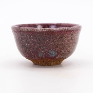 Può includere: Una piccola ciotola in ceramica fatta a mano con una superficie macchiata e strutturata. La ciotola presenta un colore bordeaux intenso con sfumature di verde acqua e una base marrone. La ciotola viene probabilmente utilizzata per servire o esporre.