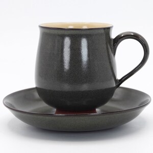 May include: A dark grey ceramic teacup and saucer set. The cup has a rounded shape and a handle. The saucer is round and has a slightly raised edge.