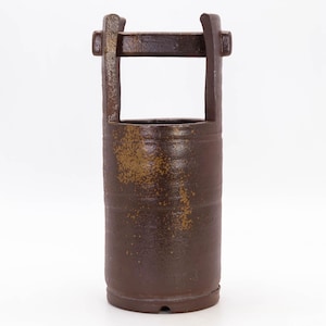 May include: A dark brown ceramic vase with a rectangular handle. The cylindrical body has a textured surface with lighter brown accents. The vase is designed with a rustic aesthetic, suitable for home decor. The height is approximately 20 cm.