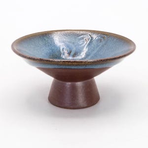Peut inclure: Un bol en céramique avec un socle. L'intérieur du bol est bleu clair et l'extérieur est marron foncé. Le bord est marron plus foncé. Le bol est sur une surface blanche.