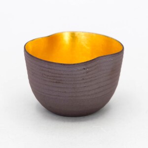 May include: A small, dark brown ceramic cup with a gold interior. The cup has a wavy rim and a textured exterior.