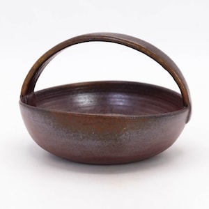 May include: A handmade, rustic ceramic bowl with a curved handle. The bowl is a deep reddish-brown colour with a smooth, slightly textured surface. The handle is a darker shade of brown, and the bowl has a simple, elegant design.