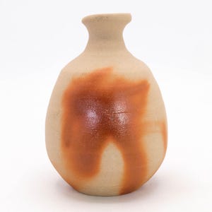 May include: A handmade ceramic vase with a rounded body and a narrow neck. The vase is a light beige color with a large, abstract, reddish-brown design on the front. The vase is likely used for decorative purposes.