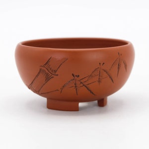 Puede incluir: Una pequeña taza de té de color terracota, de forma redondeada y con tres patas cortas. La taza presenta un diseño intrincado de bambú y libélulas tallados en la superficie.