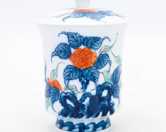 May include: A white porcelain teacup and lid with a blue and orange floral design. The cup has a wide base and a rounded body. The lid has a knob on top.