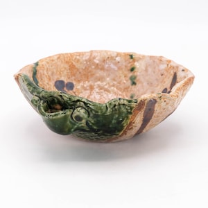 May include: A handmade ceramic bowl with a unique design. The bowl features a textured, light brown interior with dark blue and green accents. The rim is a dark green colour with a sculpted design. The bowl is approximately 15 cm in diameter.