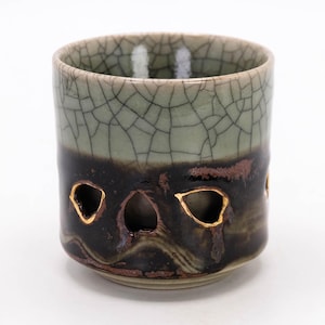 Puede incluir: Una taza de cerámica con un diseño único. La mitad superior es de color verde claro con un patrón agrietado, que pasa a una base marrón oscuro. Los detalles dorados resaltan las formas recortadas cerca de la parte inferior. La taza es probablemente hecha a mano.