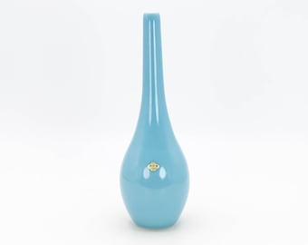 May include: A light blue ceramic vase with a narrow neck and a rounded base. The vase has a small, gold label on the front.
