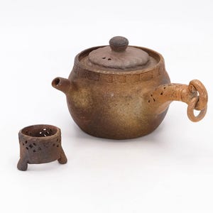 May include: A handcrafted brown ceramic teapot with a matching lid and a small, round tea warmer. The teapot features a curved handle with decorative cutouts and a spout. The tea warmer has three small legs and decorative cutouts.