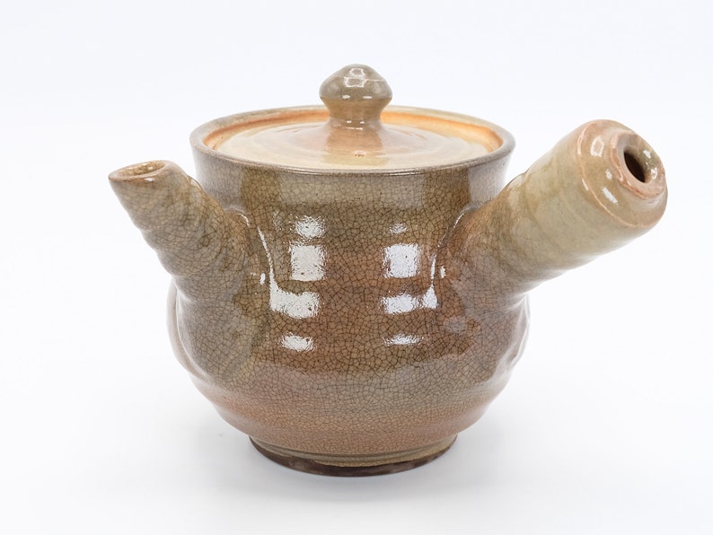 Może przedstawiać: Brązowy ceramiczny czajnik z pękniętą glazurą i dzi&oacute;bkiem z boku. Czajnik ma pokrywkę z małym uchwytem na g&oacute;rze.