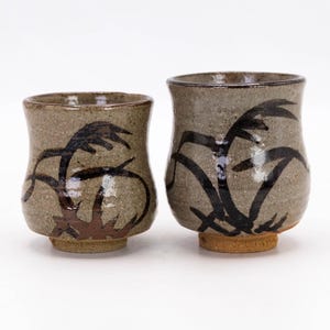 May include: Two ceramic cups with a gray glaze and black brushstroke designs of stylized grass. The cups are slightly different sizes.