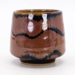 Könnte beinhalten: Eine handgefertigte Keramiktasse mit braunem Boden und einem dunkelbraunen und schwarzen Design. Die Tasse hat eine weite Öffnung und eine leicht konische Form. Der Innenraum ist dunkler braun. Die Tasse wird wahrscheinlich zum Trinken von Getränken verwendet.