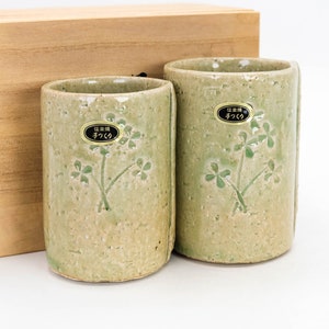 Könnte beinhalten: Zwei grüne Keramikbecher mit einem Kleeblatt-Design und einem goldenen Etikett mit der Aufschrift "伝統  手づくり" in Japanisch. Die Becher stehen auf einer Holzoberfläche.