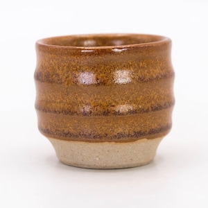 Può includere: Una piccola tazza in ceramica fatta a mano con un design strutturato e scanalato. La tazza presenta una calda glassa marrone sulla parte superiore e una base beige naturale non smaltata. La tazza viene probabilmente utilizzata per bere bevande.