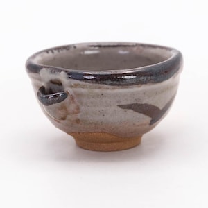 May include: A small, handmade ceramic bowl with a unique, asymmetrical shape. The bowl features a speckled gray and brown glaze with dark brown accents and a small handle. The base is a natural, unglazed brown.