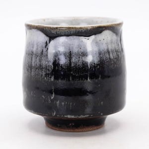 May include: A ceramic tea cup featuring a glossy black glaze on the lower portion and a textured white drip glaze near the rim. The cup has a rounded body, a slightly flared opening, and a visible unglazed brown clay base at the bottom.