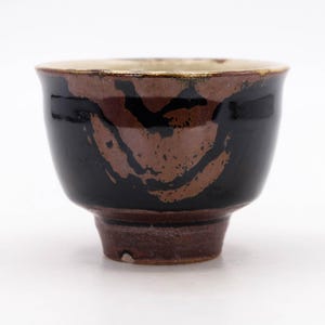 Pode incluir: Uma pequena chávena de chá em cerâmica com um vidrado preto brilhante e um padrão abstrato de pinceladas castanhas texturizadas. A chávena tem um rebordo largo e arredondado que afunila para uma base espessa de cor castanho-avermelhada.