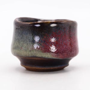 Puede incluir: Una pequeña taza de cerámica hecha a mano con un esmalte único. La taza presenta una mezcla de colores, incluyendo rojo intenso, verde y marrón oscuro. El exterior tiene una apariencia texturizada y la taza tiene una forma redondeada con un borde ligeramente ensanchado. La taza mide aproximadamente 5 cm de alto.