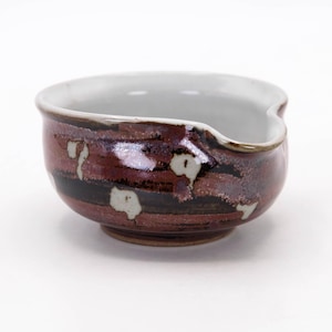 May include: A handcrafted ceramic bowl with an unusual, organic form. The bowl has a dark red and brown glaze with black and white details. The interior is light grey. The bowl has a small spout and is suitable for serving sauces or dips.
