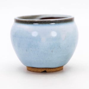 May include: A small, round ceramic pot with a light blue glaze. The rim is dark brown, and the base is a natural, unglazed clay color. The pot has a smooth, glossy finish and is suitable for small plants or decorative use.