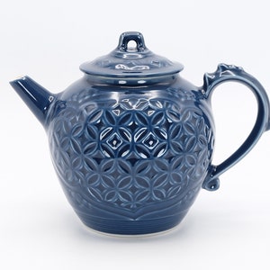 Może przedstawiać: Niebieski ceramiczny czajnik z ozdobnym, wytłoczonym wzorem. Czajnik ma pokrywkę i uchwyt.