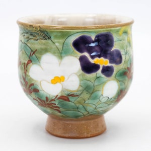 Puede incluir: Una taza de té pequeña, redonda y de cerámica con un esmalte verde y diseños florales. La taza presenta flores blancas y moradas con centros amarillos, y hojas marrones y verdes. La taza tiene una base marrón.