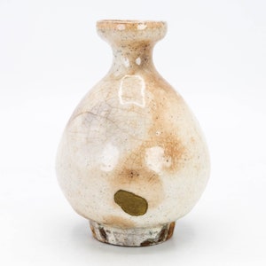 May include: A small, off-white ceramic vase with a bulbous body and a narrow neck. The vase has a speckled appearance with brown and tan accents and a crackled surface. A small, gold-coloured sticker is affixed to the side.