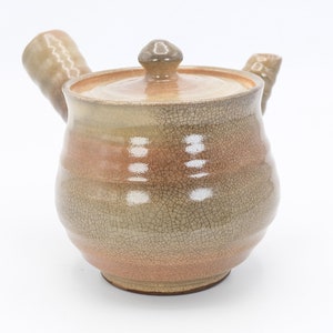 Może przedstawiać: Ceramiczny czajnik z pękniętą glazurą w odcieniach brązu i beżu. Czajnik ma zaokrąglony korpus i pokrywkę z małym uchwytem. Dzi&oacute;bek i uchwyt są lekko zakrzywione.