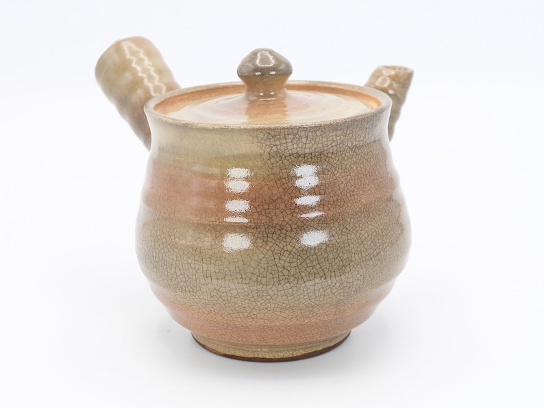 Może przedstawiać: Ceramiczny czajnik z pękniętą glazurą w odcieniach brązu i beżu. Czajnik ma zaokrąglony korpus i pokrywkę z małym uchwytem. Dzi&oacute;bek i uchwyt są lekko zakrzywione.