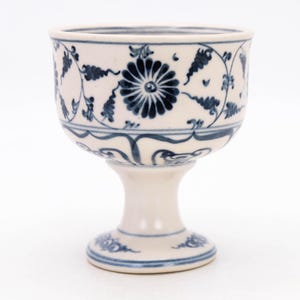 May include: A white ceramic goblet with a pedestal base, adorned with intricate blue floral patterns. The goblet features a wide bowl and a detailed design around the rim and base, showcasing a classic aesthetic.