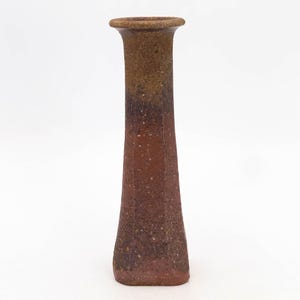 Puede incluir: Un jarrón de cerámica alto y texturizado con una parte superior ensanchada y un cuerpo hexagonal. El jarrón presenta un degradado de colores, que va del marrón oscuro en la parte superior al marrón rojizo en la base. Pequeñas motas blancas añaden detalle.