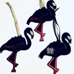 May include: Three black flamingo ornaments with red legs and white beak accents. One features the name JERRY in red, one displays the year 2026 in red, and one has a white graphic symbol. Each includes a ribbon loop for hanging purposes.