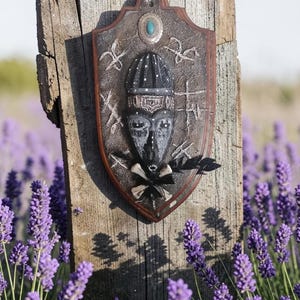 Op de afbeelding: Een decoratief schild met een tribaal maskerontwerp, gemonteerd op een verweerde houten paal. Het schild heeft een bruine rand, een centraal masker met een zwarte hoofdtooi en een turquoise stenen accent. De achtergrond toont een veld met paarse lavendel.