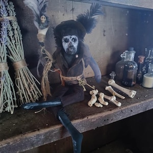 Puede incluir: Una muñeca de temática oscura con rostro pintado de blanco, maquillaje negro en los ojos y un sombrero adornado con plumas negras. La muñeca viste una chaqueta marrón, sostiene un bastón con una calavera y tiene una llave dorada en la cintura.