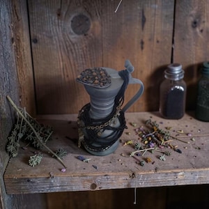 Puede incluir: Una jarra de cerámica gris mate con tapa abatible que presenta un elemento decorativo marrón circular y texturizado. La jarra está envuelta en múltiples hilos de cordón negro y cadenas metálicas finas, creando una estética oscura.