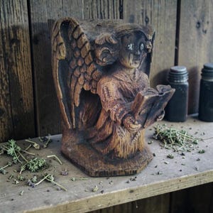 Peut inclure: Une figurine d'ange en bois sculpté avec des ailes détaillées et des robes fluides, tenant un livre ouvert. La figure présente des textures de grain de bois sombre et patiné, avec des accents de peinture blanche sur les yeux et le visage.